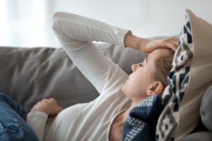 Headaches may be symptoms of more serious underlying medical conditions. In this image, a young woman in pain is lying on a couch with her hand on her forehead.